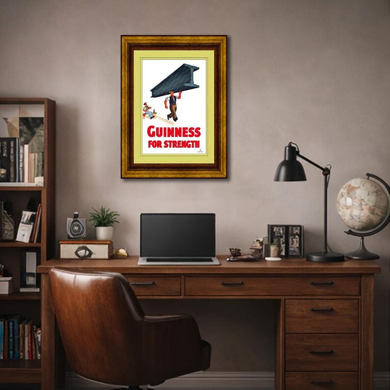 May include: Framed vintage Guinness advertisement. The artwork features a man carrying a large steel beam, with the text "Guinness for Strength" in red. The frame is a dark wood with a gold inner border. The image is displayed on a wall above a wooden desk.