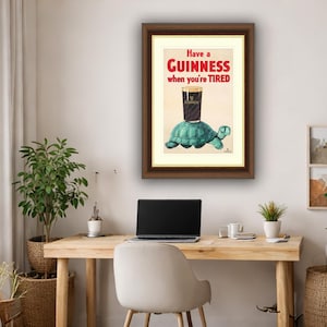 K&ouml;nnte beinhalten: Gerahmter Druck mit einer Guinness-Werbung. Es zeigt ein Pint Guinness, das auf einer t&uuml;rkisfarbenen Schildkr&ouml;te balanciert, mit dem Text "Have a Guinness when you're TIRED" in Rot und Schwarz.