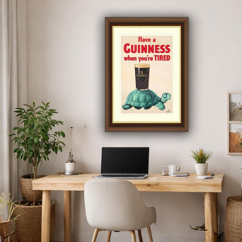 K&ouml;nnte beinhalten: Gerahmter Druck mit einer Guinness-Werbung. Es zeigt ein Pint Guinness, das auf einer t&uuml;rkisfarbenen Schildkr&ouml;te balanciert, mit dem Text "Have a Guinness when you're TIRED" in Rot und Schwarz.
