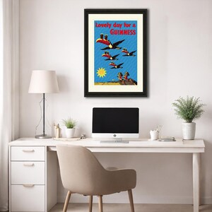 May include: Framed vintage Guinness advertisement featuring toucans carrying pints of Guinness against a blue background with the text "Lovely day for a Guinness." The artwork includes a yellow sun and figures at the bottom.