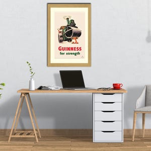 Puede incluir: Un anuncio vintage enmarcado de Guinness, con un rodillo de vapor y el texto "Guinness for strength" en rojo. El p&oacute;ster est&aacute; encima de un escritorio de madera con un port&aacute;til, una taza roja y un mueble de cajones blanco.