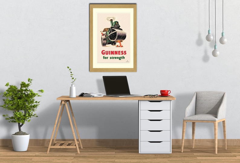 Puede incluir: Un anuncio vintage enmarcado de Guinness, con un rodillo de vapor y el texto "Guinness for strength" en rojo. El p&oacute;ster est&aacute; encima de un escritorio de madera con un port&aacute;til, una taza roja y un mueble de cajones blanco.