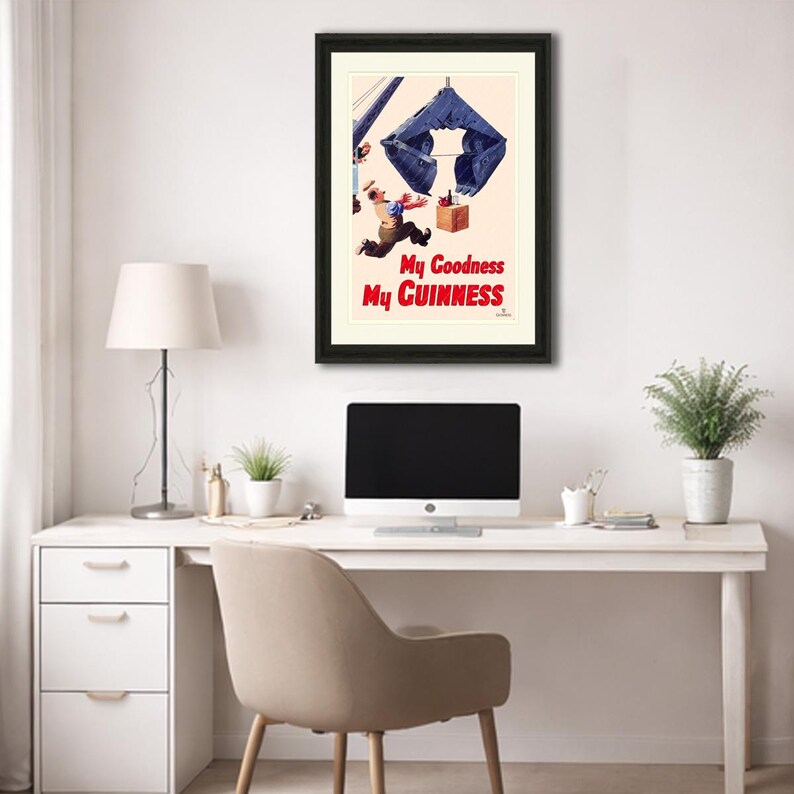 Pode incluir: An&uacute;ncio vintage emoldurado da Guinness. A obra de arte retrata um homem caindo com um grande objeto azul acima dele. O texto "My Goodness My Guinness" est&aacute; em vermelho. A impress&atilde;o emoldurada &eacute; exibida em uma mesa branca.
