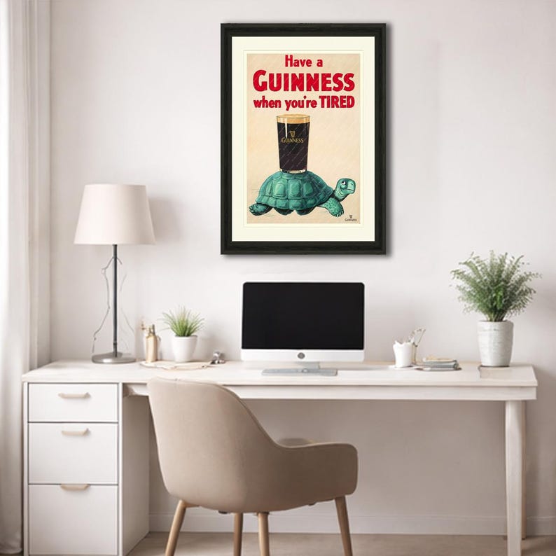 K&ouml;nnte beinhalten: Gerahmter Kunstdruck mit einer skurrilen Illustration. Ein Pint Guinness-Bier ruht auf einer t&uuml;rkisfarbenen Schildkr&ouml;te, mit dem Text "Have a Guinness when you're TIRED" in Rot und Schwarz. Das Kunstwerk ist schwarz gerahmt und vor einem cremefarbenen Hintergrund.