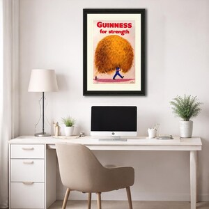 May include: Framed vintage Guinness advertisement. The artwork features a man carrying a large, golden-brown bundle with the text "GUINNESS for strength" in red. The framed print is displayed on a white desk with a computer and lamp.
