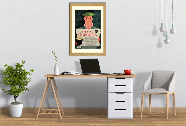 May include: A framed vintage advertisement for Guinness stout. The artwork depicts a man in a green cap reading a newspaper with the headline "Evening Guinness." The image is set in a home office with a desk, chair, and a potted plant.