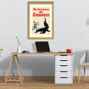 May include: Framed vintage Guinness advertisement featuring a seal balancing a pint of Guinness. The poster has the text "My Goodness My Guinness" in red and white. The poster is in a gold frame, displayed above a wooden desk with a laptop and a red mug.