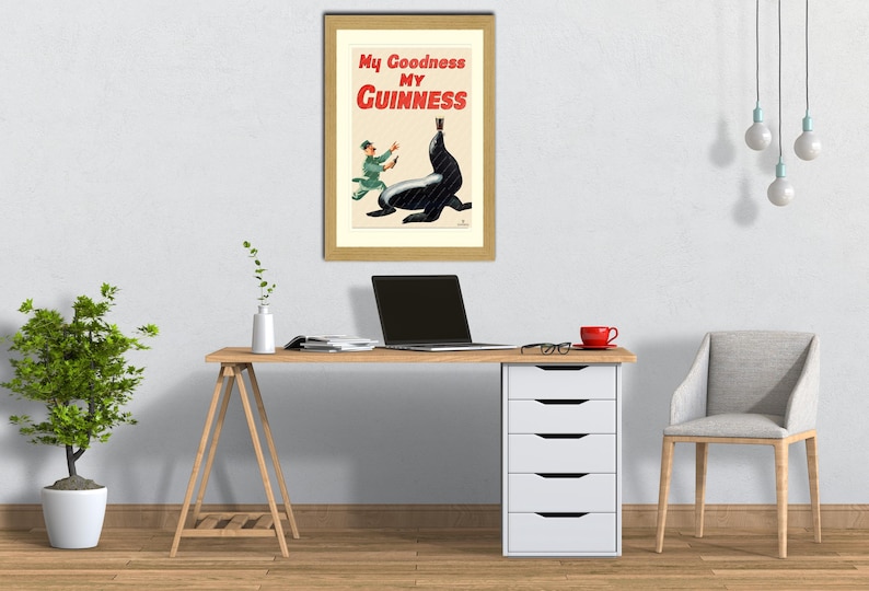May include: Framed vintage Guinness advertisement featuring a seal balancing a pint of Guinness. The poster has the text "My Goodness My Guinness" in red and white. The poster is in a gold frame, displayed above a wooden desk with a laptop and a red mug.
