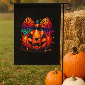 May include: A black Halloween garden flag with a smiling jack-o'-lantern and a large bow. The bow has a leopard print design with orange, purple, and blue accents. Several pumpkins of different sizes and colours are in the foreground.