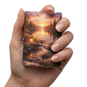 May include: A rectangular art print depicts a sunset over a river with a lantern in the foreground. The artwork features a vibrant sky with orange and yellow hues, a calm body of water, and lush green trees. The print is held in a hand.