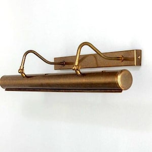 May include: A bronze-colored picture light fixture with a rectangular backplate and a curved arm design. The light has a cylindrical body and is mounted on a white wall. The fixture is designed to illuminate artwork.