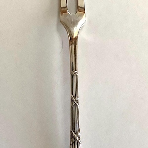 May include: Silver serving fork with two tines. The handle is decorated with vertical lines, cross-shaped designs, and a bow-shaped element. The fork is designed for serving food.