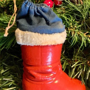 May include: A festive Christmas ornament shaped like a red boot with a blue fabric top and white trim. The boot is made of a glossy material and has a loop for hanging. The ornament is set against a backdrop of green pine needles and red berries.