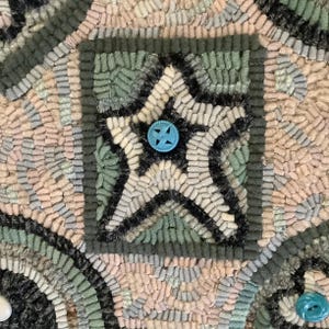 May include: Close-up of a handmade rug with a star design. The star is made of light blue and white yarn and has a turquoise button in the center. The rug is made of various shades of blue, green, and pink yarn.