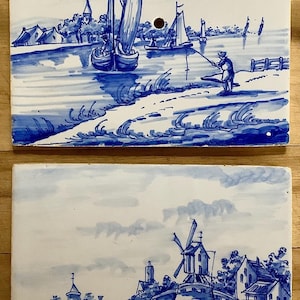 May include: Two blue and white ceramic tiles with Dutch landscape scenes. One tile depicts sailboats on a river, and the other shows a windmill and buildings. Each tile has a hole for hanging.