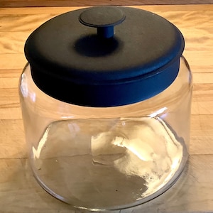 May include: A clear glass storage jar with a wide cylindrical body and a smooth, matte black lid. The lid features a centered, circular knob handle on top, providing a simple and functional design for this transparent kitchen container.