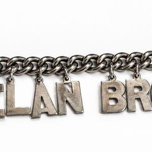 May include: A silver-toned metal charm bracelet featuring individual letter charms that spell out CLEVELAN BROWNS, with a small football-shaped charm attached to the end of the thick curb chain link structure.