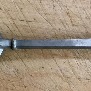 May include: A vintage metal spoon with a rounded bowl and a long, slender handle. The handle has the text "DS PATER" engraved on it. The spoon is made of a dull, silver-colored metal and rests on a wooden surface.