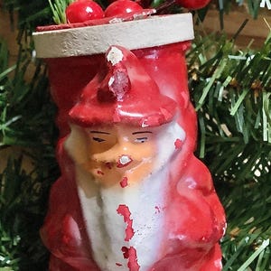 May include: A vintage Christmas ornament shaped like Santa Claus, painted red with white accents. The ornament has a weathered appearance, with visible paint chipping. Red berries and green foliage are in the background.