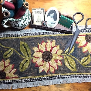 May include: A hand-stitched rug with a sunflower design in shades of yellow, green, and red. The rug has a black and white checkered border and fringe. A pair of antique scissors and a spool of thread are in the background.