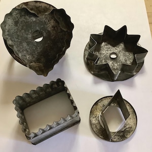 May include: Four vintage metal cookie cutters in various shapes: a heart, a starburst, a scalloped rectangle, and a diamond within a circle. The cutters have a rustic, aged appearance with signs of wear.