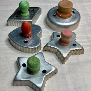 May include: A set of five metal cookie cutters with wooden handles in various shapes including a diamond, a circle, a heart, and two stars. The handles are painted in green, orange, and pink, while the cutters feature fluted edges.