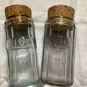 May include: Two vintage glass spice jars with cork lids. The jars are clear glass with a ribbed design. The jar on the left is labeled "ALL SPICE" and the jar on the right is labeled "PEPPER".