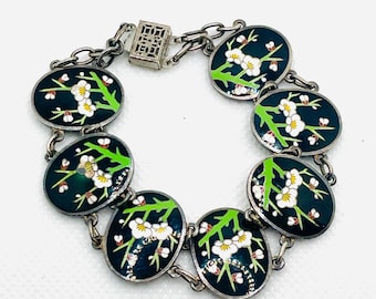 Op de afbeelding: Zilverkleurige armband met ovale schakels met een donkerblauwe achtergrond. Elke schakel toont een wit bloemmotief met groene stelen. De armband heeft een decoratieve sluiting.