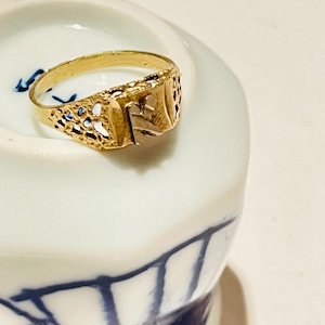 Op de afbeelding: Een goudkleurige ring met een sierlijk ontwerp. De ring heeft een vierkant middengedeelte met de letter "E" en decoratieve uitsparingen langs de band. Hij ligt op een wit en blauw keramisch oppervlak.