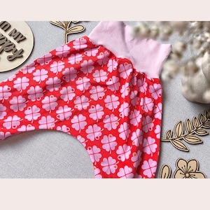 Könnte beinhalten: Rote Babyhose mit einem rosa Kleeblattmuster. Der Bund ist hellrosa. Das Kleeblattmuster zeigt rosa vierblättrige Kleeblätter auf rotem Hintergrund. Die Hose ist aus weichem Stoff.
