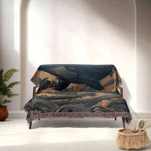 May include: A navy blue and gold throw blanket featuring a whale design. The blanket depicts a whale swimming in the ocean with waves. The throw has a fringed edge and is draped over a wooden sofa.