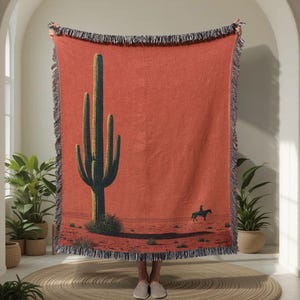 May include: A woven throw blanket in a terracotta orange hue featuring a large green saguaro cactus on the left and a small silhouette of a person riding a horse on the right. The blanket has a decorative fringe border along all four edges.