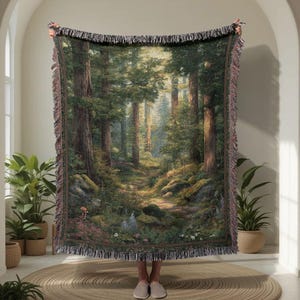 May include: A woven tapestry blanket featuring a forest scene with tall trees, a path, and sunlight. The blanket has a fringed edge and is held up by a person. The color palette includes greens, browns, and beige.