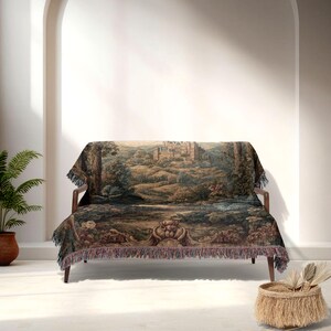 May include: A tapestry throw blanket draped over a wooden sofa. The blanket features a detailed landscape scene with a castle, trees, and water in shades of green, brown, and blue. The blanket has a fringed edge.