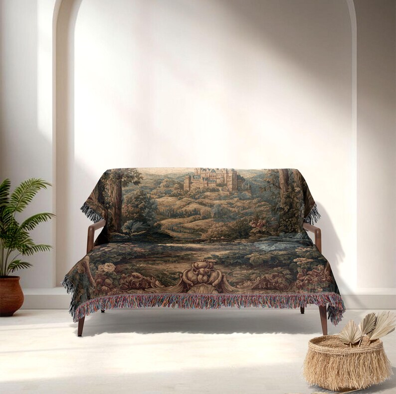 May include: A tapestry throw blanket draped over a wooden sofa. The blanket features a detailed landscape scene with a castle, trees, and water in shades of green, brown, and blue. The blanket has a fringed edge.
