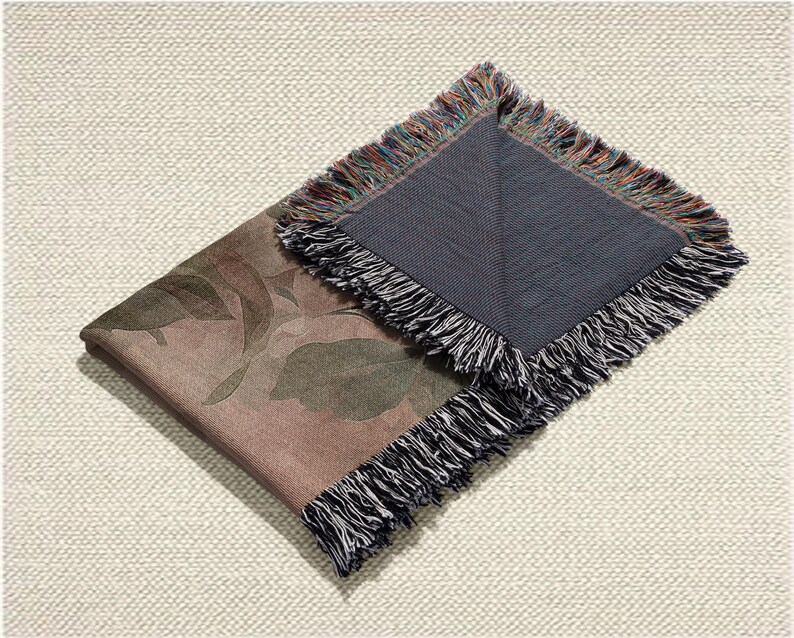 May include: Folded throw blanket with a botanical leaf pattern in shades of brown, green, and grey. The blanket has a dark grey underside and is trimmed with a multi-coloured fringe. The blanket is folded on a textured, off-white surface.