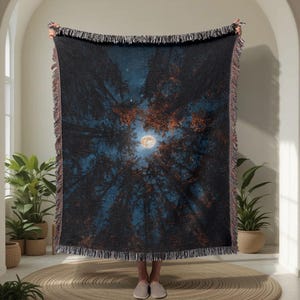 Puede incluir: Una manta tejida que presenta una escena de cielo nocturno mirando hacia arriba a través de altos pinos hacia una luna llena brillante. La manta tiene una paleta de colores azul oscuro y negro con follaje anaranjado y bordes con flecos.