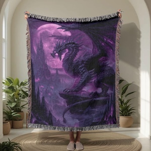 Può includere: Una coperta tessuta con un'illustrazione dettagliata di un drago nelle tonalità del viola e del nero. Il drago è appollaiato su una roccia con una luna piena sullo sfondo. La coperta ha un bordo sfrangiato.