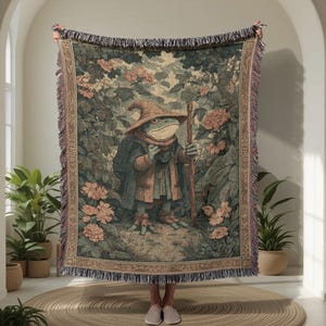 May include: Woven tapestry blanket featuring a whimsical illustration of a frog dressed as a wizard, holding a staff, set in a floral garden scene. The blanket has a fringed border and is predominantly green, brown, and beige.