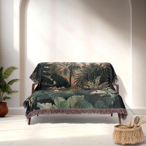 May include: A dark green and pink throw blanket with a tropical jungle print, draped over a wooden sofa. The blanket has a fringe along the bottom edge. A potted plant and a woven basket are in the foreground.