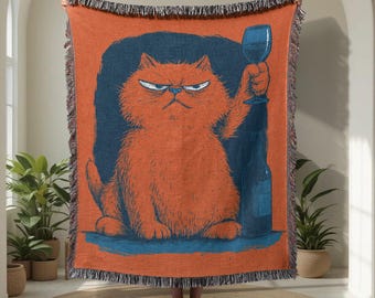 May include: Orange throw blanket with a grumpy cat illustration holding a wine glass and bottle. The cat is set against a dark blue background. The blanket has a fringed edge.