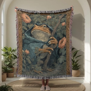 May include: Woven tapestry blanket with a fantasy illustration of a frog in a blue cloak holding a staff. The design includes peach-coloured flowers and green foliage, with a fringed border. The blanket is displayed indoors.