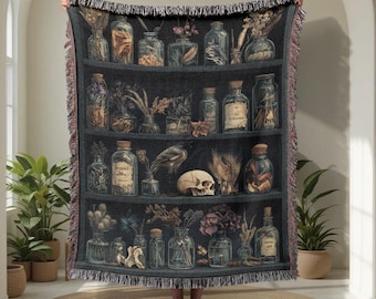 May include: A dark blue woven throw blanket featuring a print of a shelf filled with apothecary jars, dried plants, and a skull. The blanket has a fringed edge and is held up by a person.