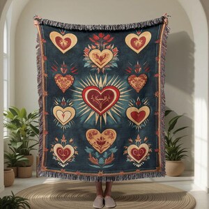 May include: A dark teal throw blanket with a repeating pattern of stylized hearts in red, gold, and cream. The central heart is surrounded by sun rays. The blanket has a fringed edge and is held up in front of a person.