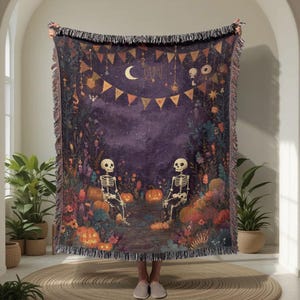 May include: A woven Halloween throw blanket featuring a whimsical scene with two skeletons, pumpkins, and a crescent moon. The blanket has a dark purple background with colourful floral and banner details, and a fringed edge.