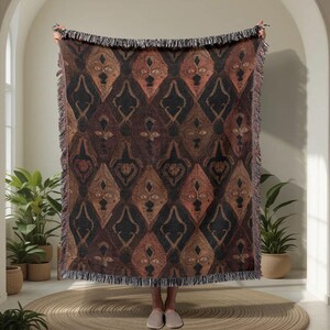 May include: A woven throw blanket with a repeating pattern of stylized faces in shades of brown, black, and rust. The blanket has a fringed edge and is held up, showcasing the design. The overall theme is bohemian and artistic.