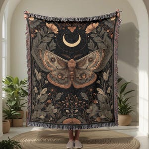 May include: A dark woven throw blanket with a moth design, a crescent moon, and floral details. The moth is in brown and tan shades against a black backdrop. The blanket has a fringed edge, and is suitable for a sofa or bed.