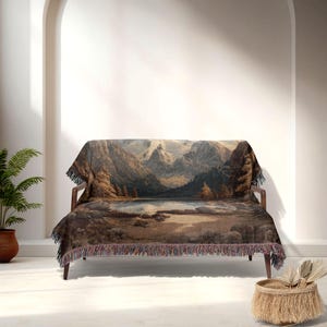 May include: A woven throw blanket draped over a wooden sofa. The blanket features a landscape scene with mountains, a lake, and trees in shades of brown, gray, and blue. The blanket has a fringed edge.