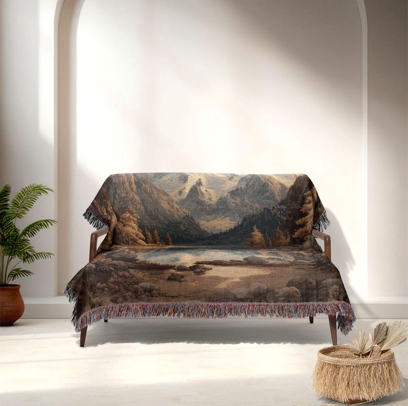 May include: A woven throw blanket draped over a wooden sofa. The blanket features a landscape scene with mountains, a lake, and trees in shades of brown, gray, and blue. The blanket has a fringed edge.