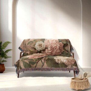 May include: A floral throw blanket on a wooden sofa. The blanket displays large pink and cream peony flowers with green leaves on a beige backdrop. The edges have a fringe trim.